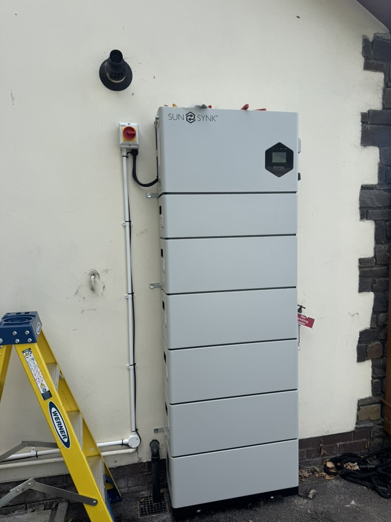 Tall Sunsynk home battery system installed against an exterior wall with an electrical isolator and neatly routed cabling